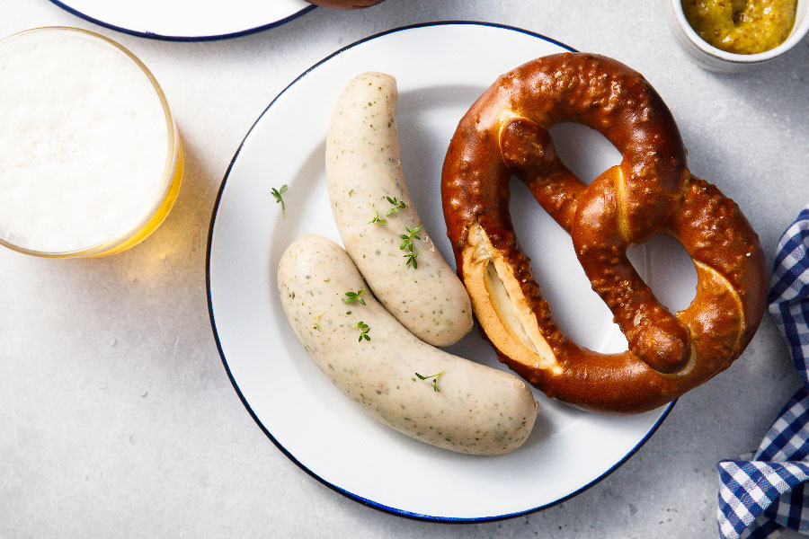 Leckere Weisswürste mit einer Breze und einem kleinen Topf it süßem Senf. Dabei steht ein Glas frisch eingeschänktes Bier.